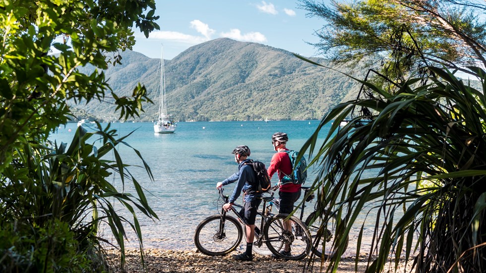 Two male bikes parked on the beach just off the Queen Charlotte Track with a yacht and the Marlborough Sounds behind.