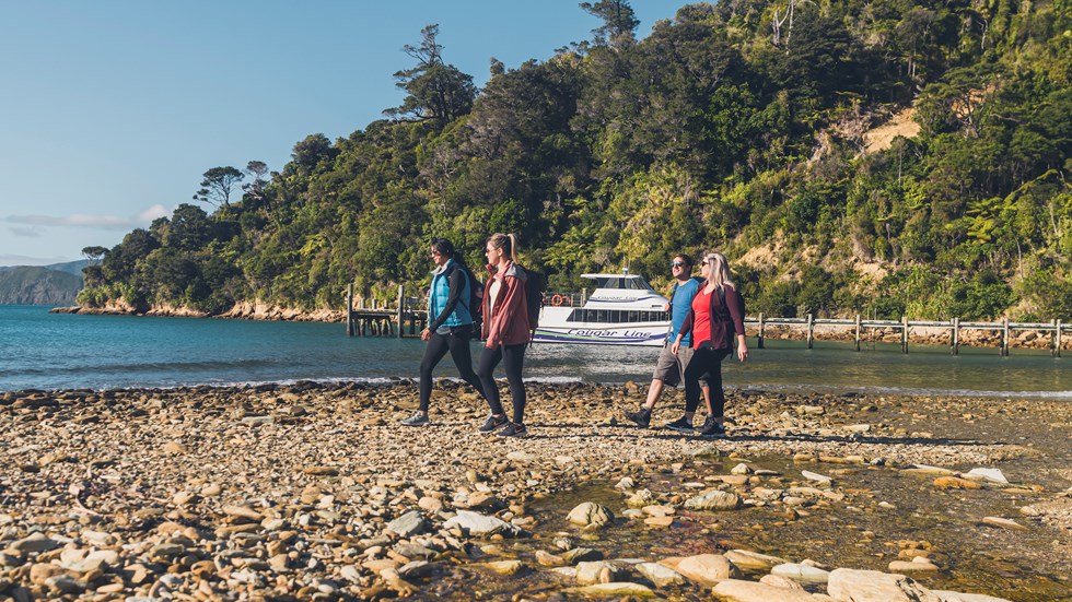 Four people walk along the beach at Ship Cove/Meretoto with a Cougar Line boat in the background.