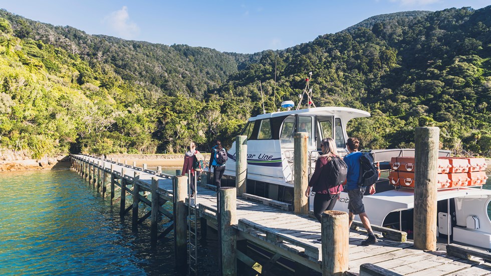 Four people walk on the jetty at Ship Cove/Meretoto next to a Cougar Line boat in the Marlborough Sounds, New Zealand.