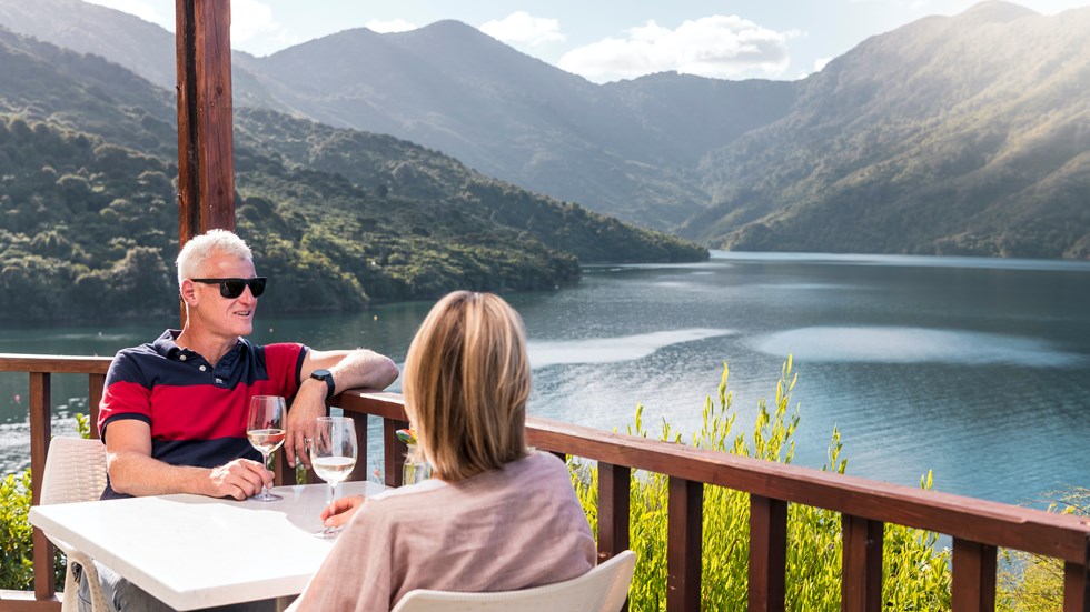 A couple sit at a table on the deck of the Punga Cove restaurant with wine, admiring the view of Endeavour Inlet in the Marlborough Sounds, New Zealand.
