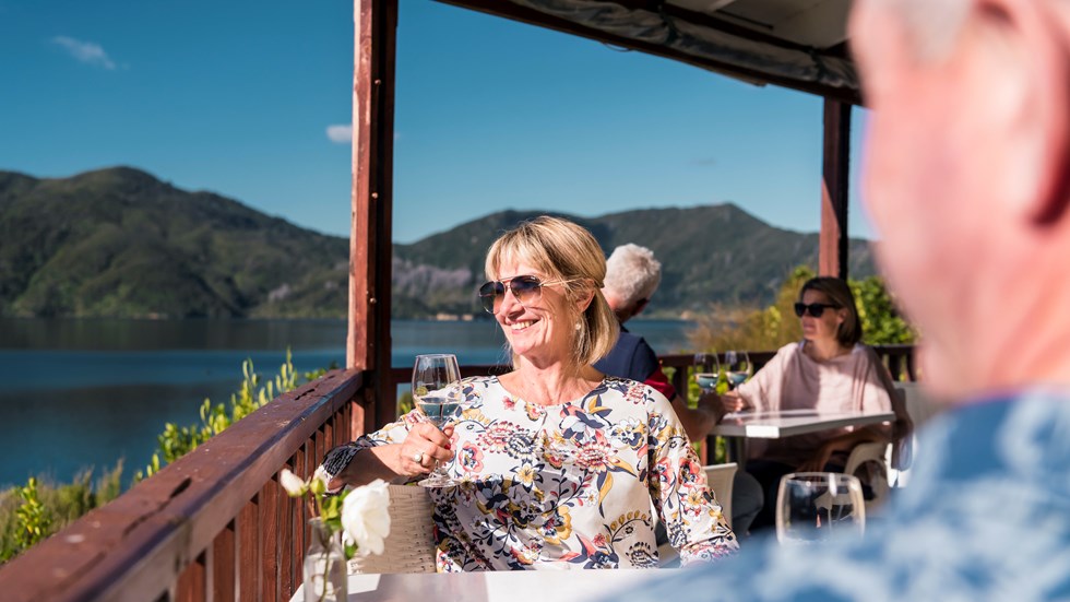A woman with glass of wine sits on balcony with partner and couple behind, and view of Marlborough Sounds at Punga Cove